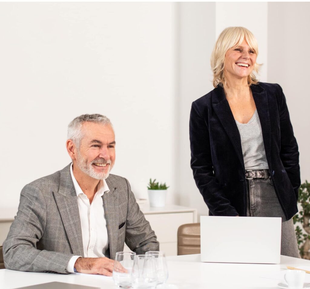 services3 Executive interim management team smiling during a business meeting in a bright office, with one leader standing beside a colleague.